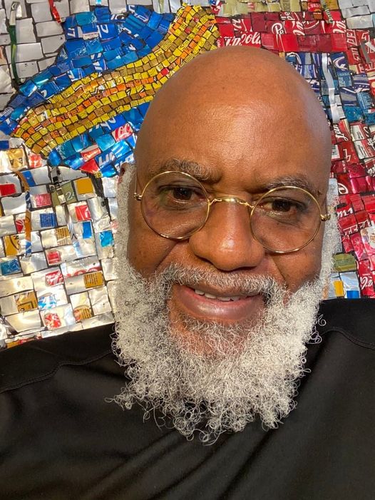 a picture showing moyo okediji wearing a pair of glasses sitting in front of his artwork