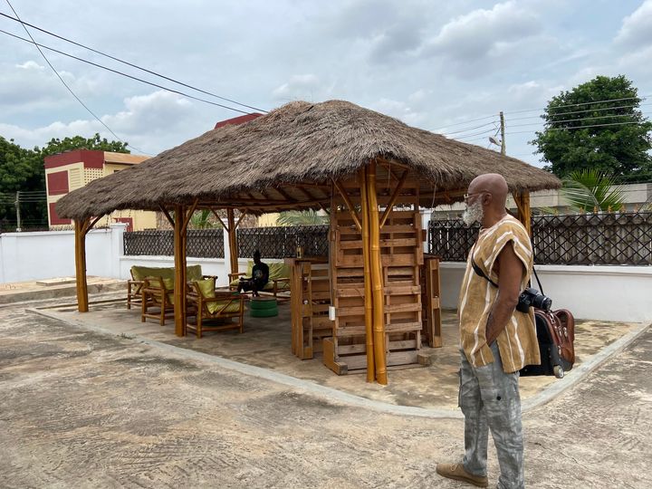 a picture showing moyo okediji standing next to a summer hut