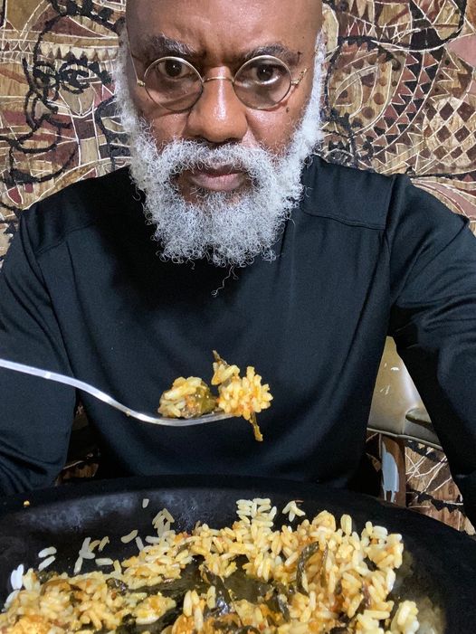 a picture showing moyo okediji eating from a plate and behind him is one of hi art piece.