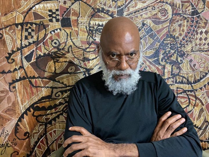 a picture showing moyo okediji sitting with his arms crosseed in front of one of his art piece